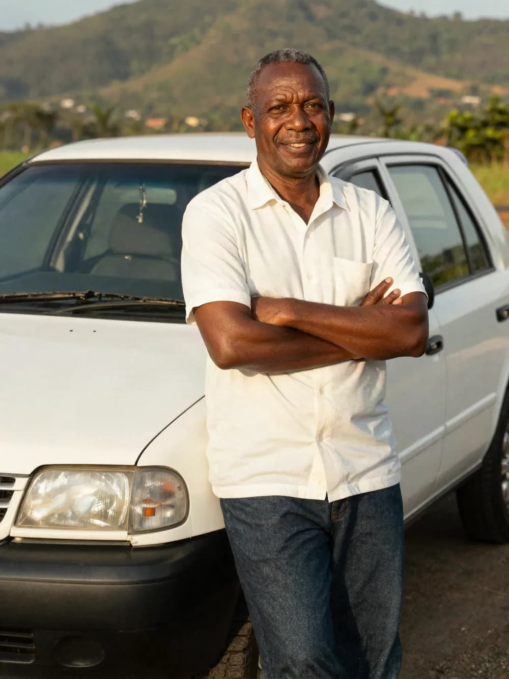 A driver-partner standing beside his white hatchback with the hills behind him