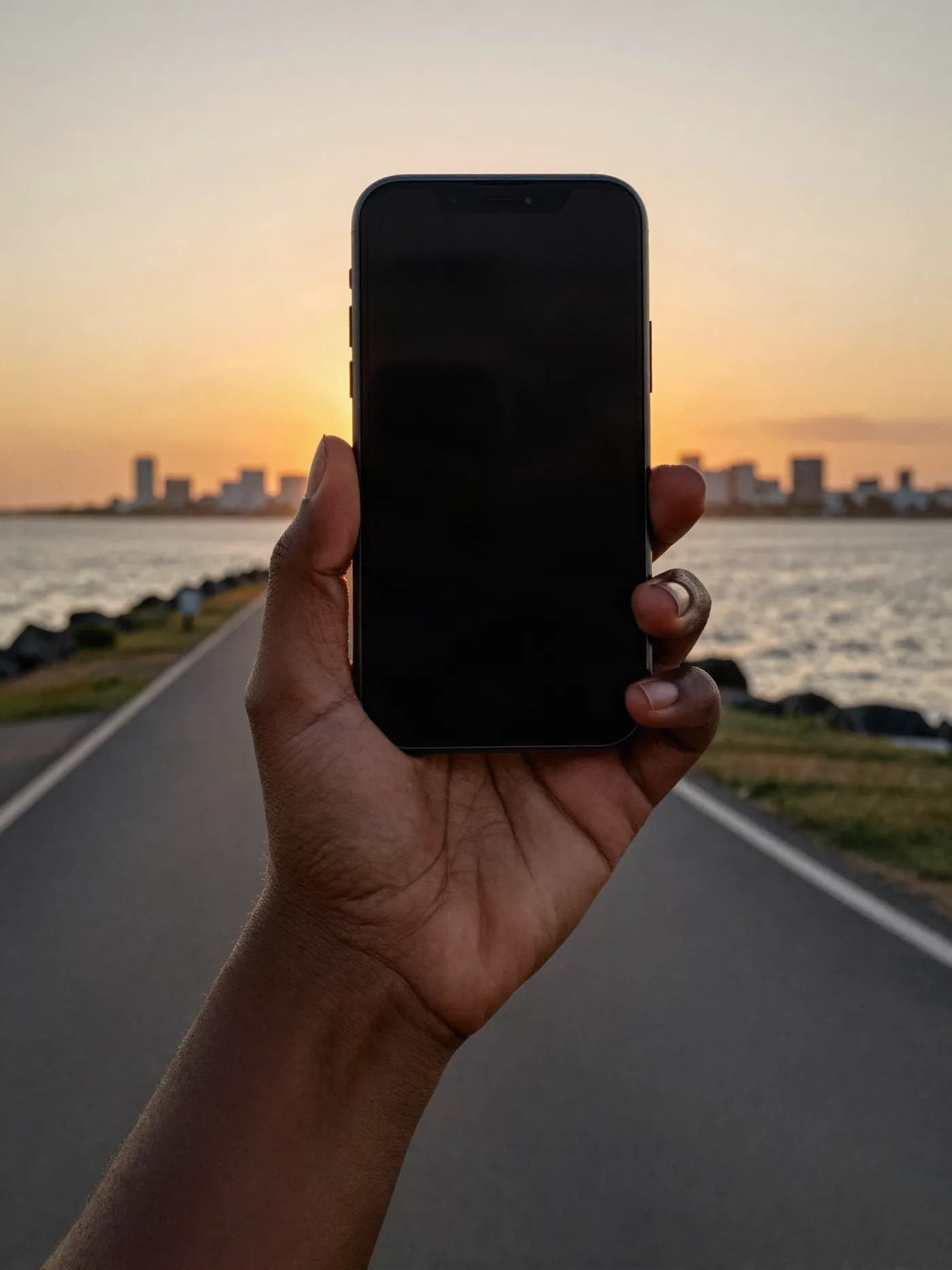 A hand holding up a phone on the Portmore causeway at sunset, sea on both sides and the city skyline behind